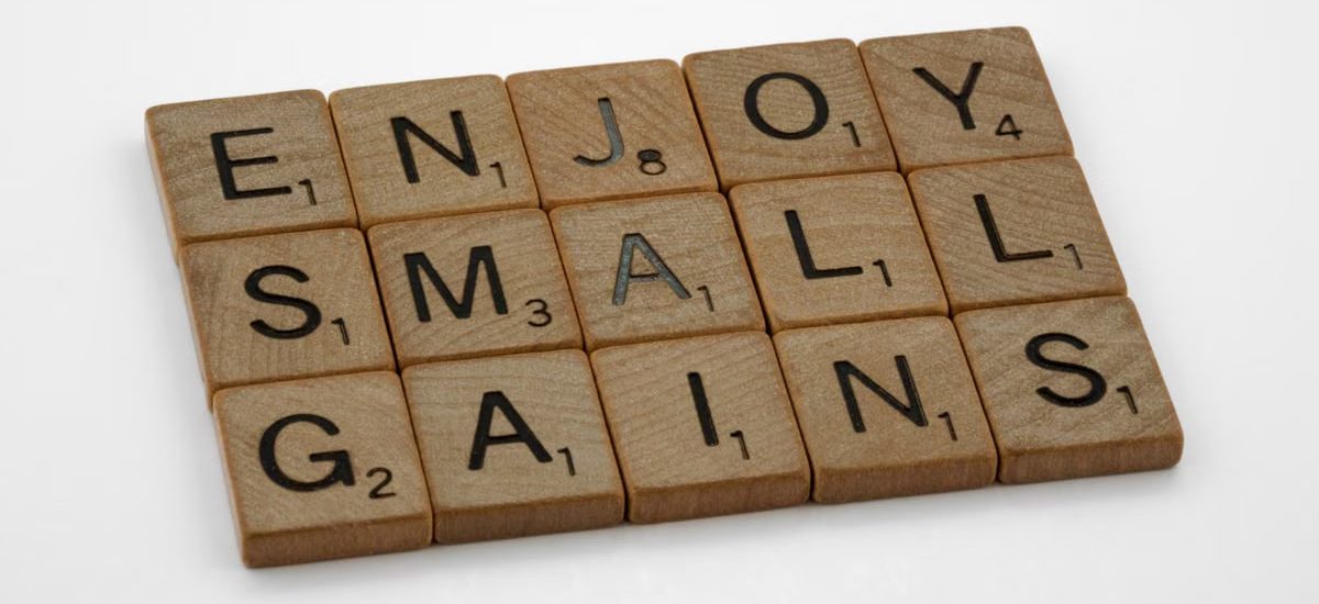 Close-up of Scrabble tiles spelling 'Enjoy Small Gains' on a white surface.